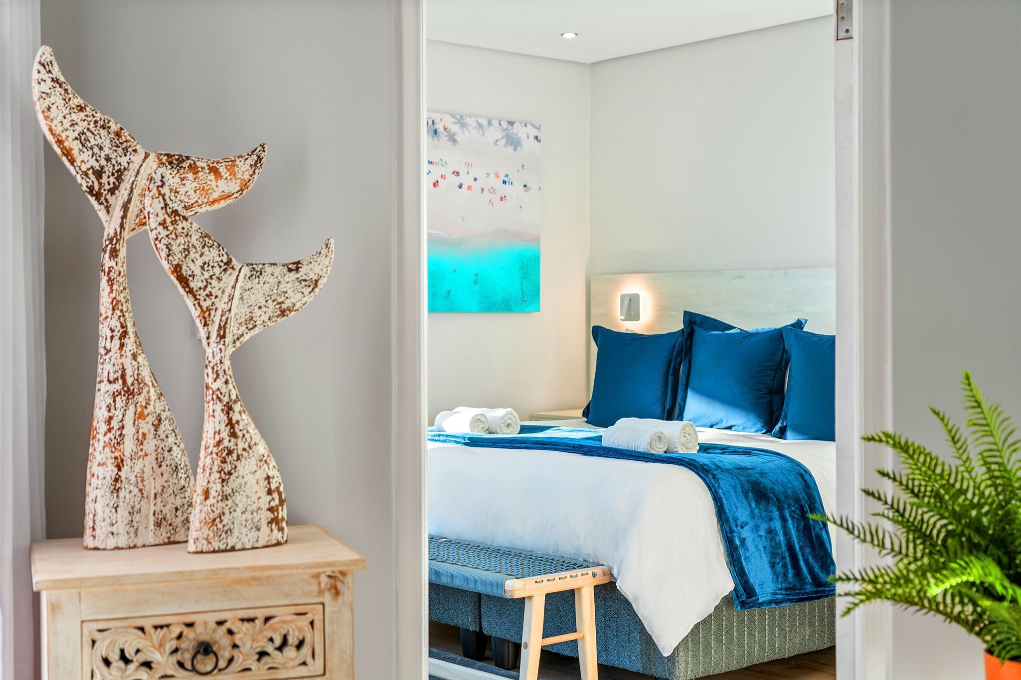 A bedroom with a bed, pillows, a bedside table, a wall art piece, and a plant.