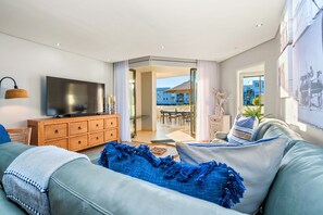 Ein modernes Wohnzimmer mit Flachbildfernseher, einer Holzkommode, einem blauen Sofa und einem Essbereich, der durch eine Glastür sichtbar ist.