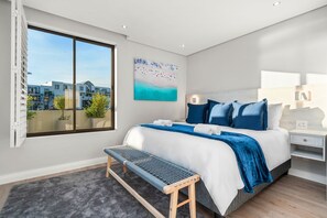 A modern bedroom with a large window, a bench, a bed with blue bedding, and a painting on the wall. - Ocean Mist 105 (Cape Town)