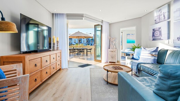 Ein modernes Wohnzimmer mit Fernseher, Holzschrank, blauem Sofa und Essbereich mit Blick ins Freie.