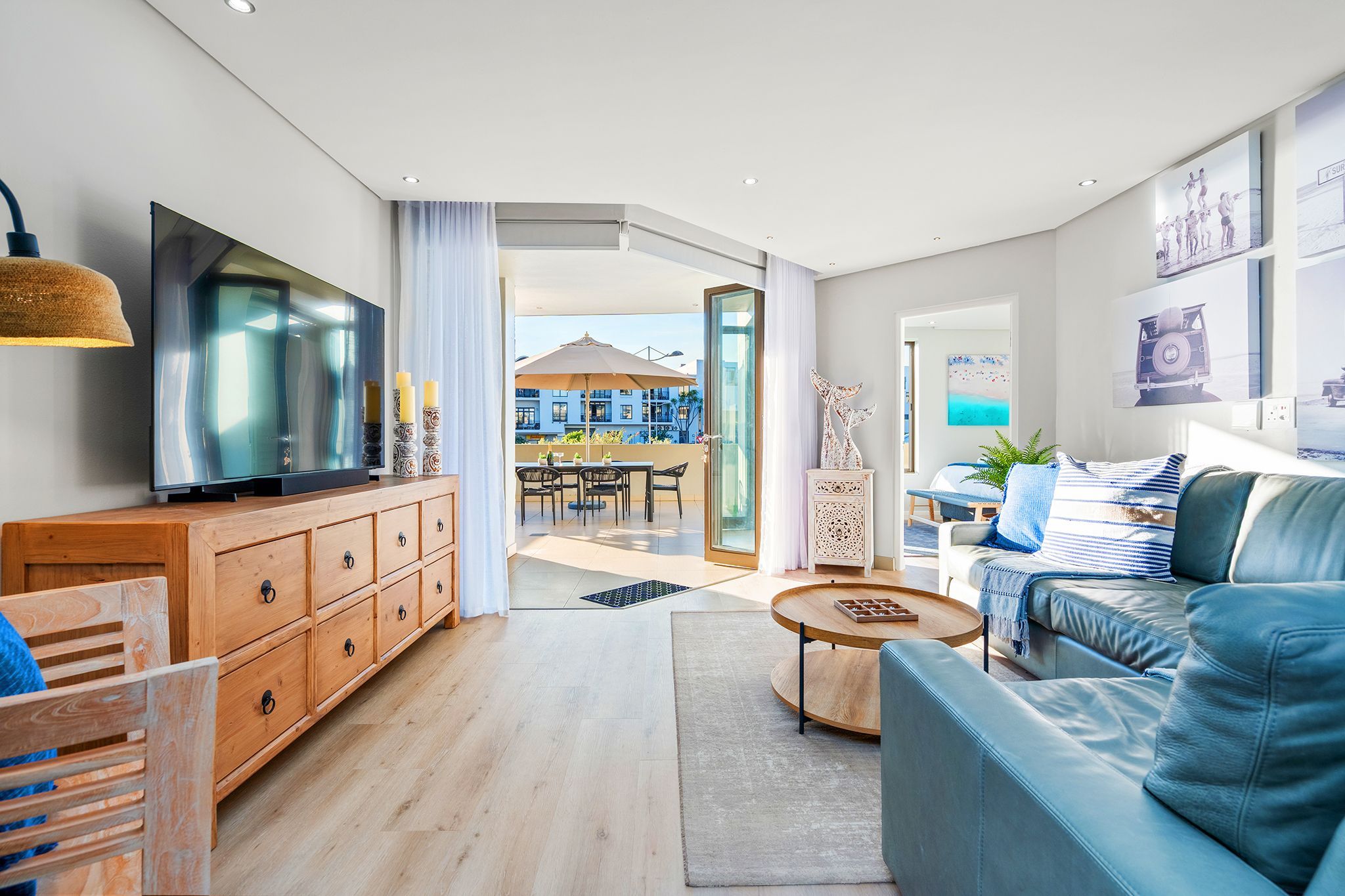 A modern living room with a TV, wooden cabinet, blue sofa, and a dining area with outdoor view.