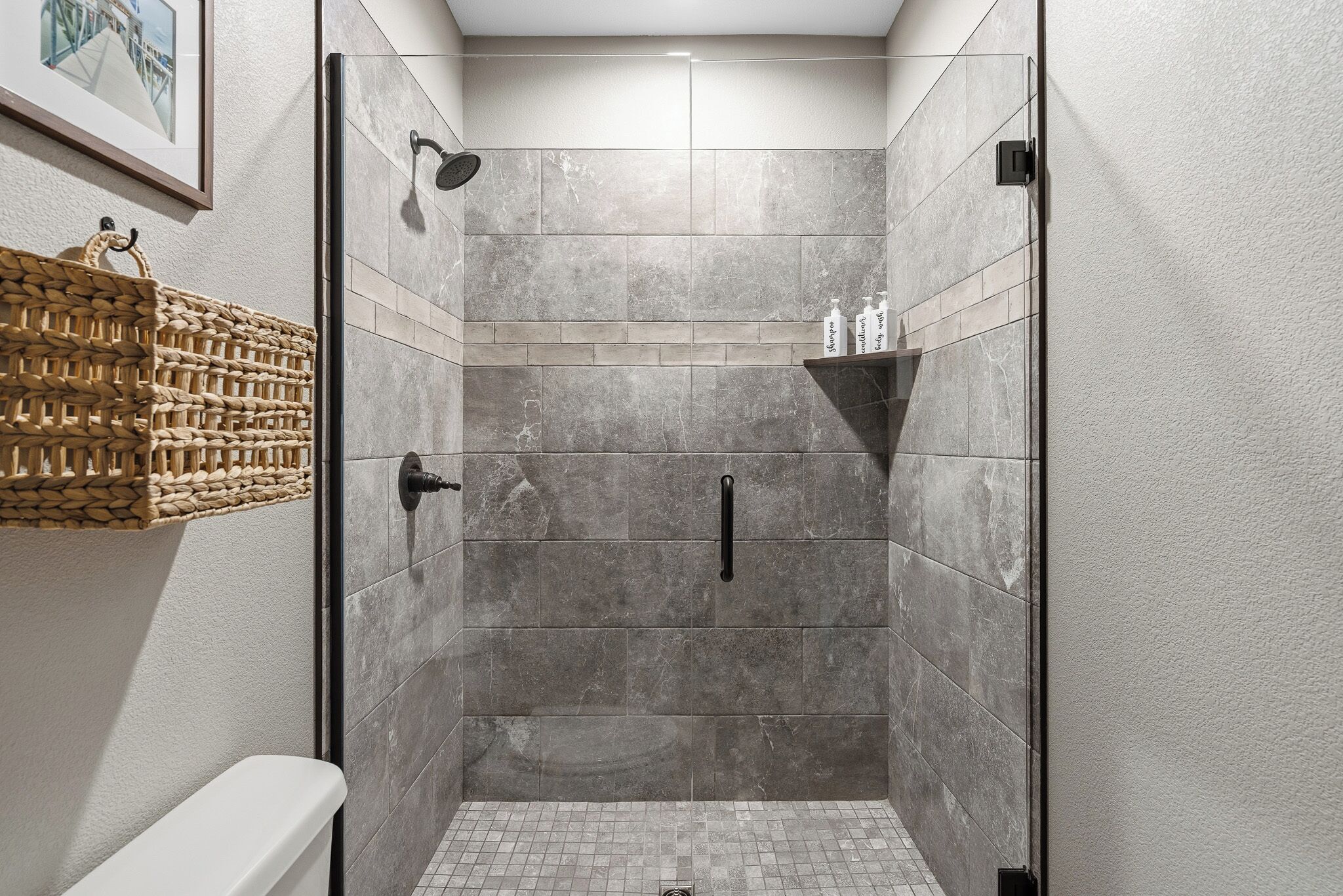 Una ducha a nivel con azulejos grises, un cabezal de ducha negro, un perchero y un estante pequeño con artículos de tocador.