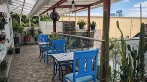 Un patio techado con sillas y mesas azules, plantas en macetas y vista de la ciudad.