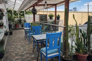 Eine überdachte Terrasse mit blauen Stühlen und Tischen, Topfpflanzen und Blick auf die Stadtlandschaft.