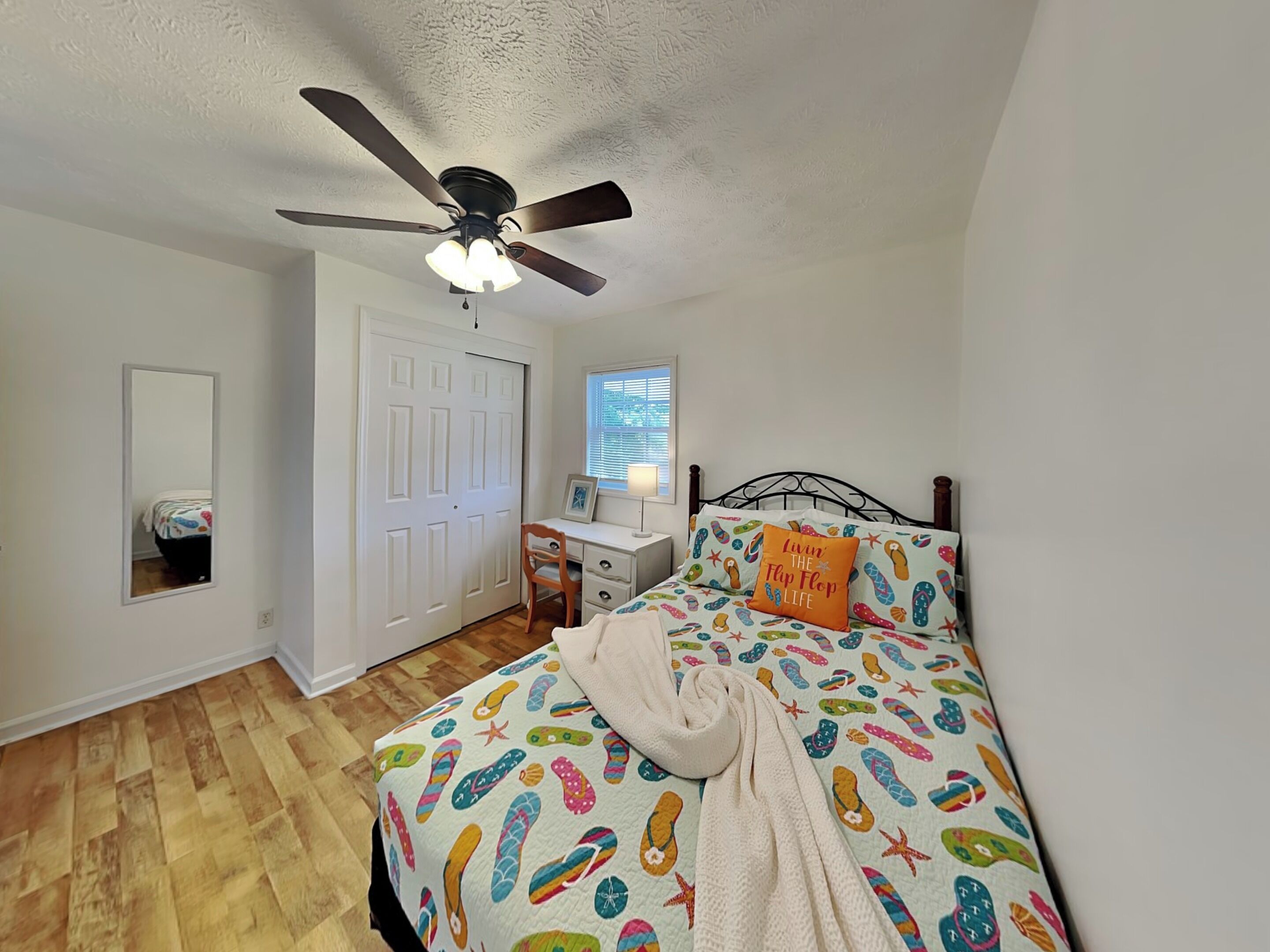 A bedroom with a bed, a desk, a chair, a ceiling fan, a window, and a door.
