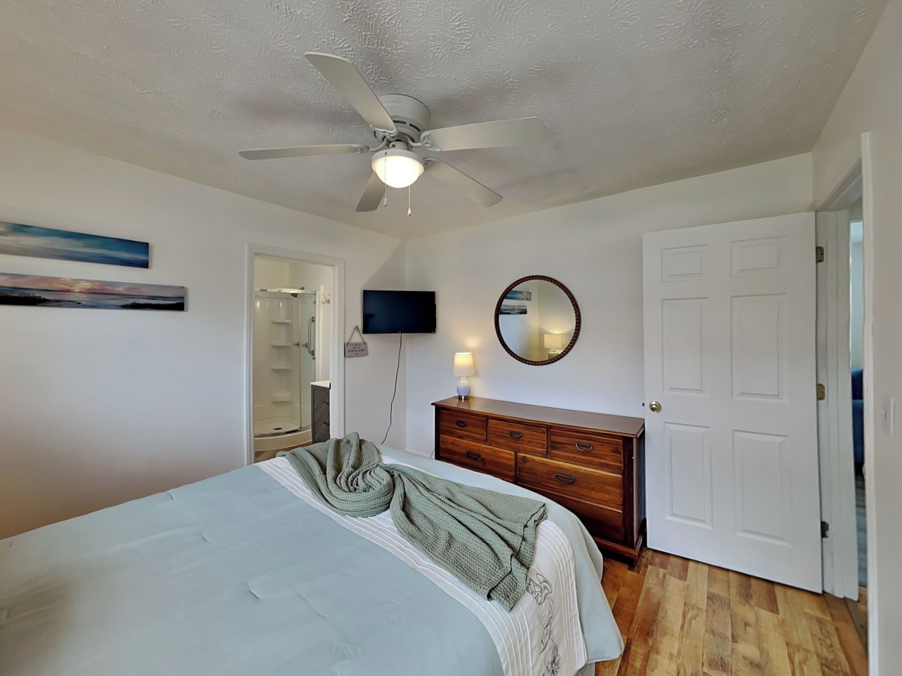 Un dormitorio con cama, ventilador de techo, televisor, espejo, cómoda y una puerta.