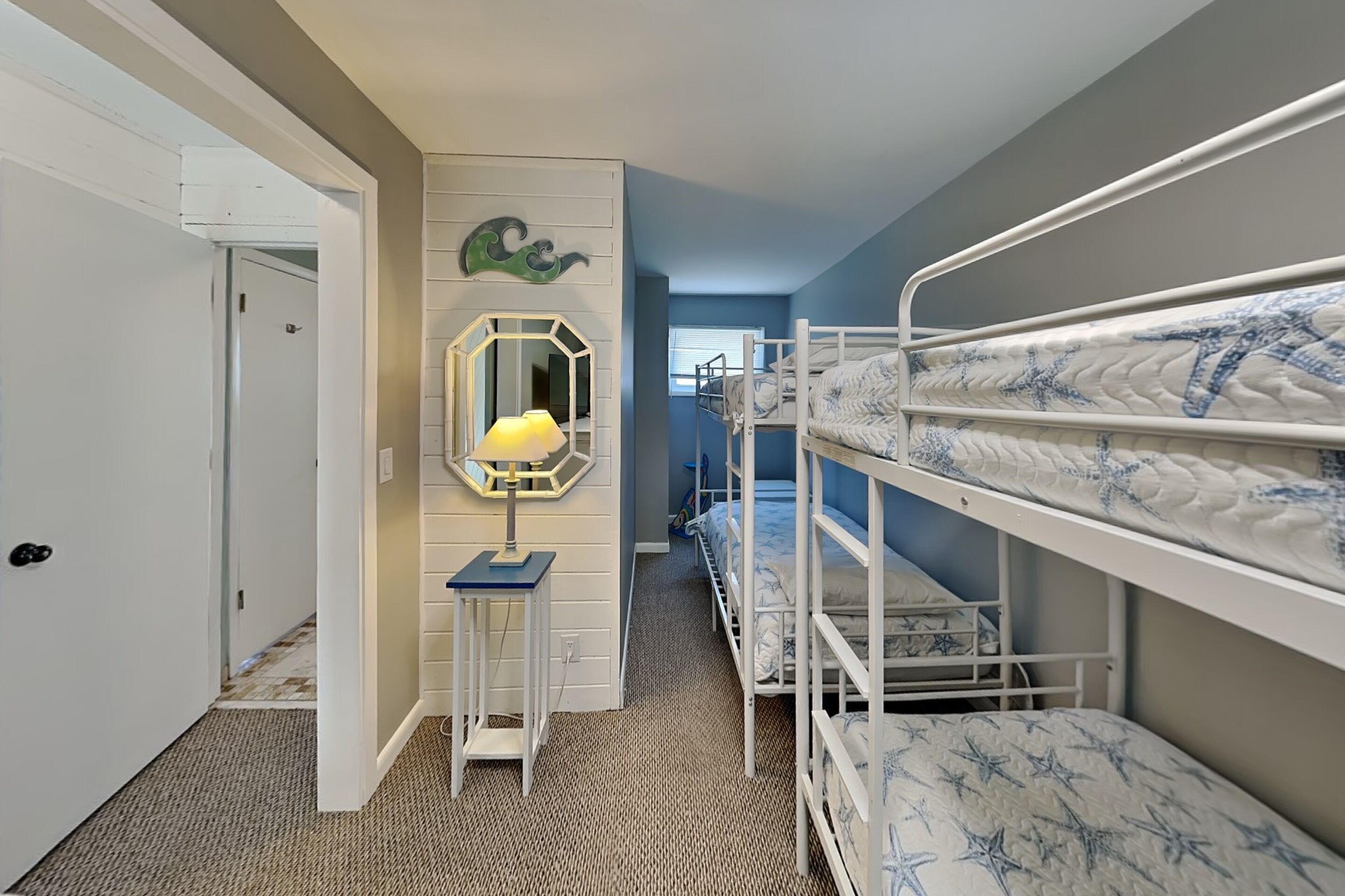 A room with bunk beds, a mirror, a lamp, and a door leading to another room.