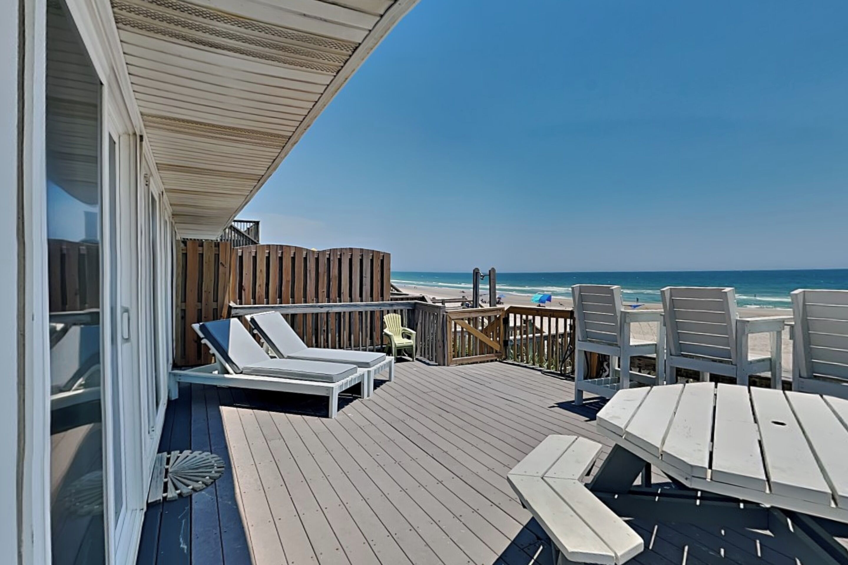 A wooden deck with a view of the beach, lounge chairs, and a table.