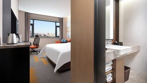 Ein Hotelzimmer mit Bett, Schreibtisch mit Wasserkocher, Stuhl, großem Fenster und Badezimmer mit Waschbecken und Handtuchhalter.