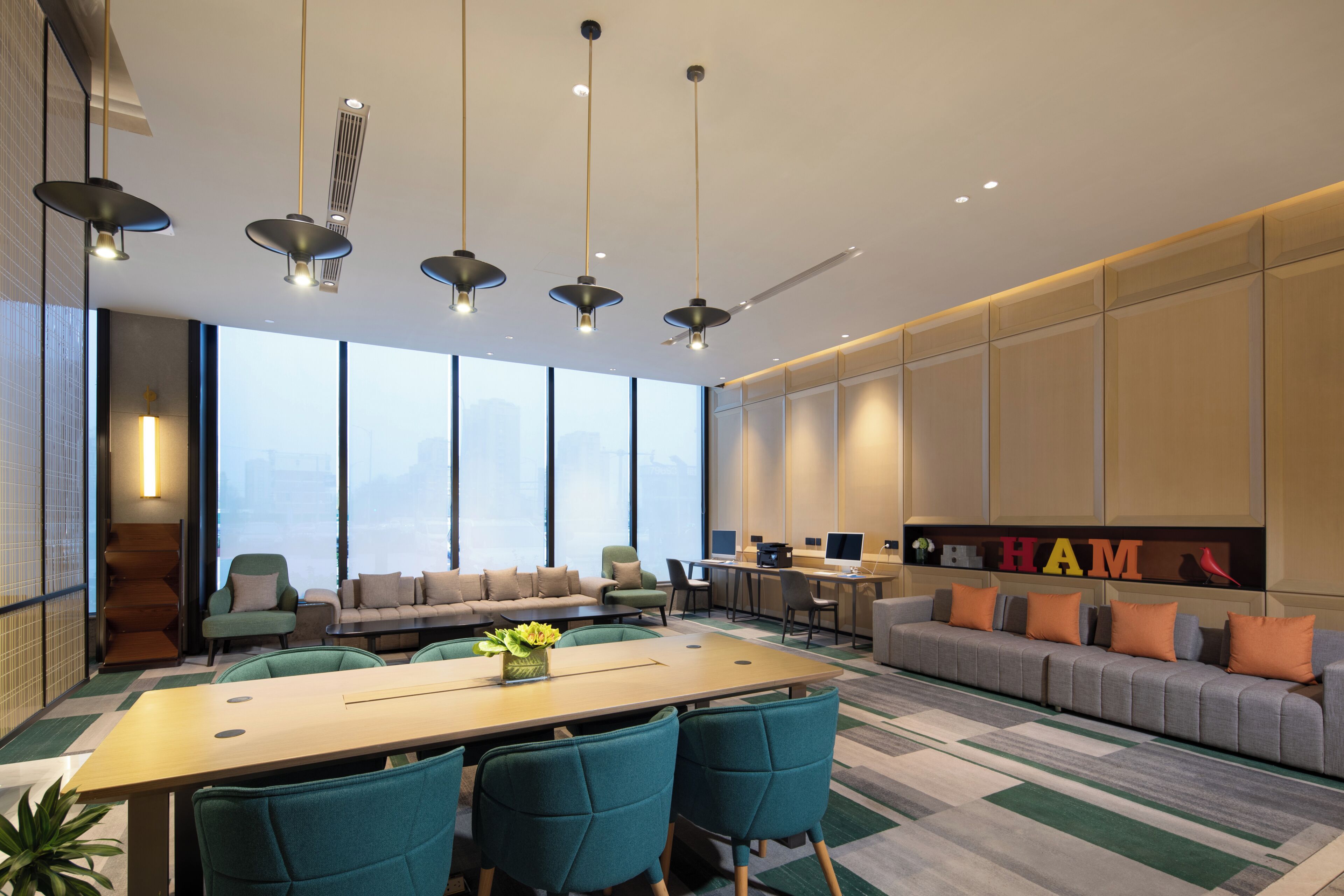 A modern conference room with a long table, teal chairs, a sofa with orange cushions, and a large window with a city view.