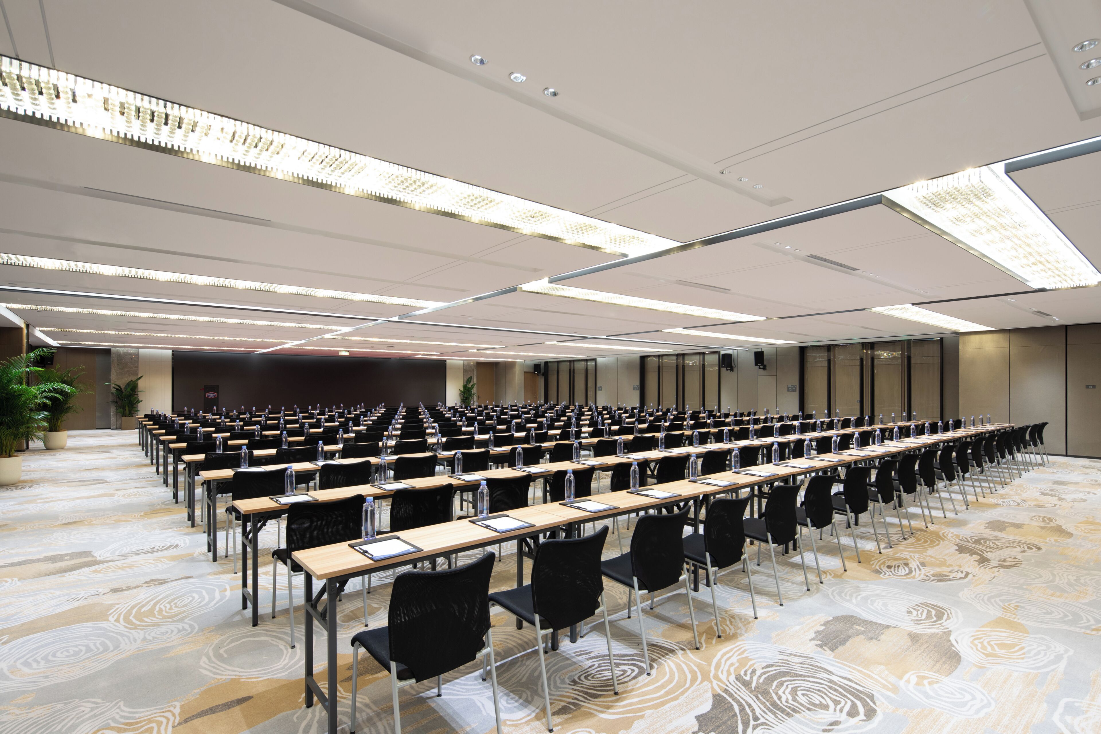 Un salón de conferencias con filas de mesas y sillas, una alfombra estampada e iluminación en el techo.