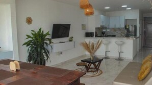 Una sala moderna con una mesa de comedor de madera, televisor, zona de cocina y área de estar con sofá y taburetes de bar.
