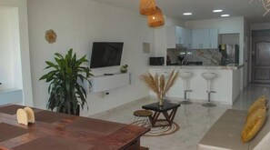 Ein modernes Wohnzimmer mit Esstisch aus Holz, einem Fernseher, einer Küchenzeile und einer Sitzecke mit einer Couch und Barhocker.
