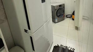 A compact kitchen with a white refrigerator, a washing machine, and a small window. - Recanto Nascer do Sol - your refuge in Cabedelo (Cabedelo)
