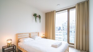 Une chambre à coucher avec un lit en bois, des lampes de chevet, une petite table, une fenêtre avec des rideaux et une vue sur les bâtiments à l’extérieur.