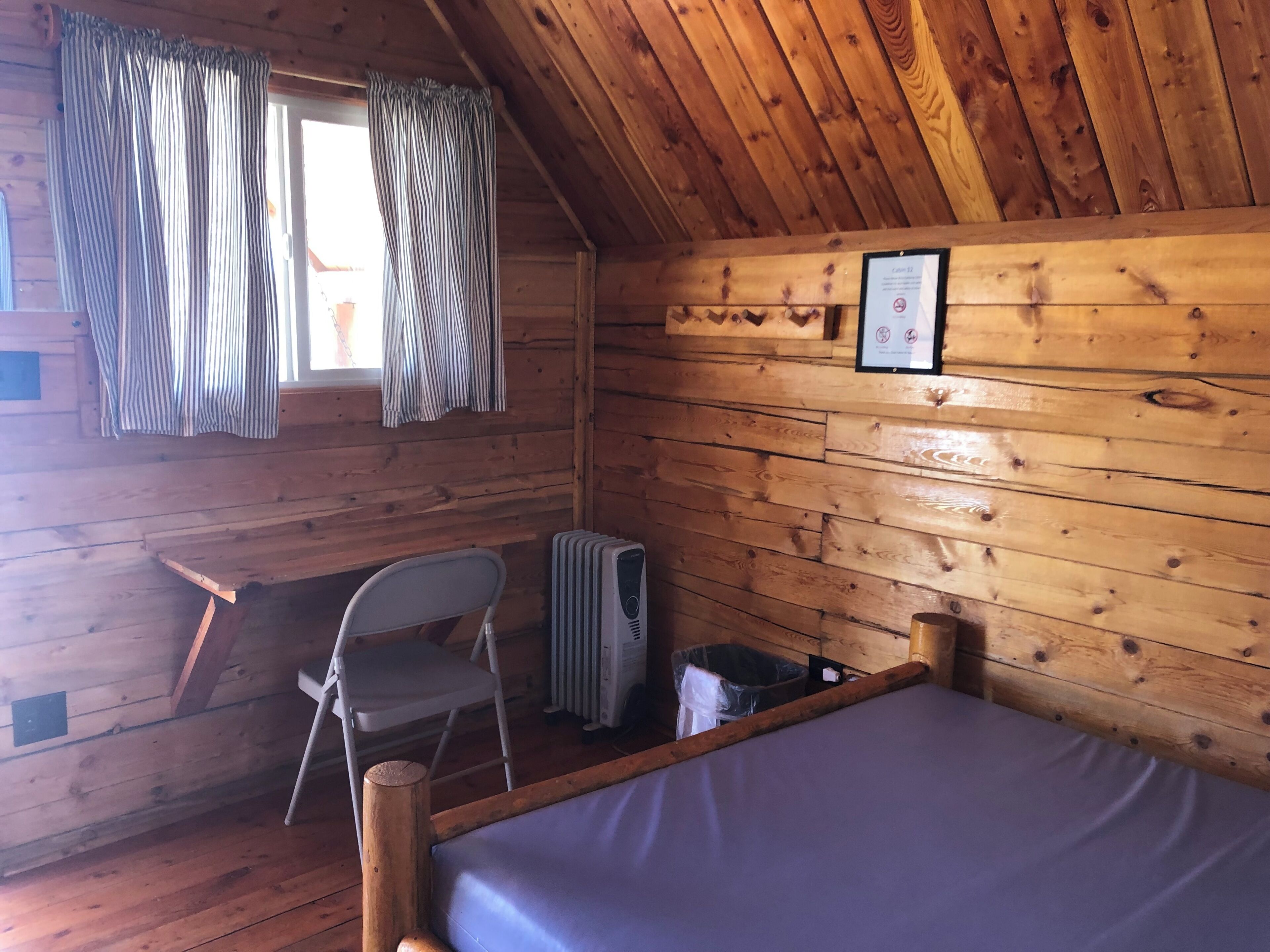 A wooden cabin room with a bed, a chair, a radiator, and a window with curtains.