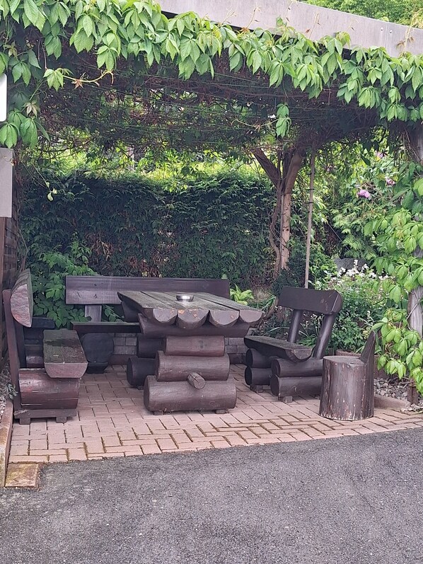 Ein rustikaler Außenbereich zum Sitzen mit Holztisch und Bänken, umgeben von Grünpflanzen und einem Pflasterweg.