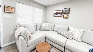 A modern living room with a sectional sofa, a round wooden coffee table, and a window with blinds.