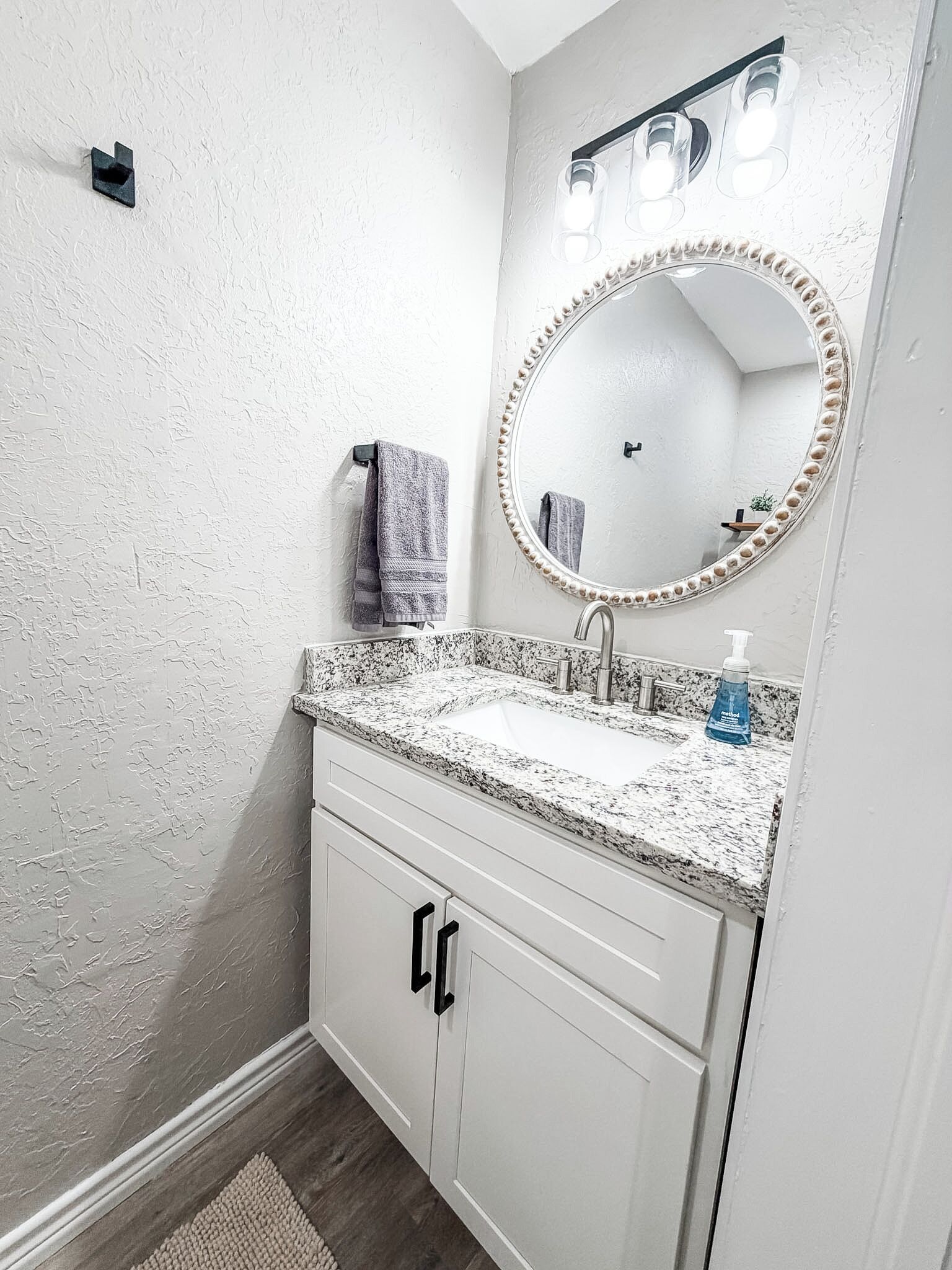 Une salle de bain avec un lavabo doté d’un plan de travail en granit, un miroir rond avec éclairage intégré et un porte-serviettes.
