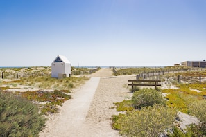 A sandy path leading to a white building with a blue roof, surrounded by vegetation and a wooden fence. - Seaside Maisonette with Stunning Views (Leucate)