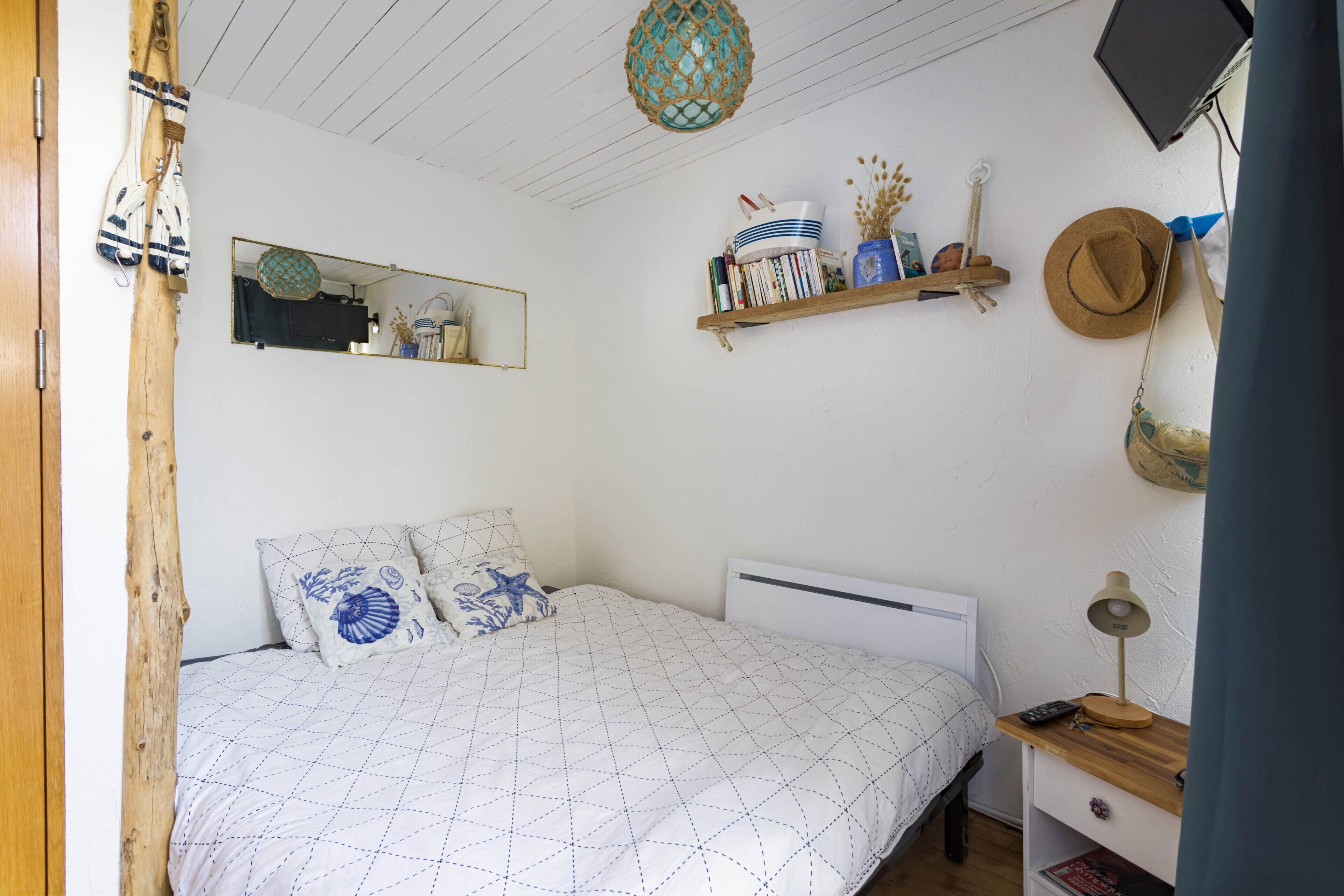 A bedroom with a bed, a bedside table with a lamp, a shelf with books and decorative items, a mirror, and a hanging hat.