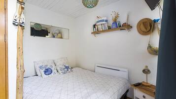 A bedroom with a bed, a bedside table with a lamp, a shelf with books and decorative items, a mirror, and a hanging hat.
