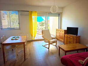 Ein Zimmer mit Esstisch, Stühlen, einer Couch, einem Fernseher auf einem Holzschrank und einer Glastür mit gelben Vorhängen.