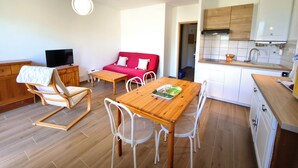 Een ruimtelijk ingerichte zithoek met een eettafel, stoelen, een rode bank, een televisie en een kitchenette.