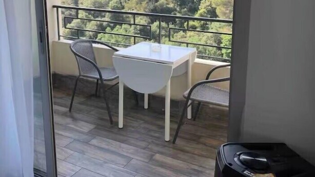 Un balcón con una mesa y sillas, un ventanal de vidrio y vista a un área boscosa.