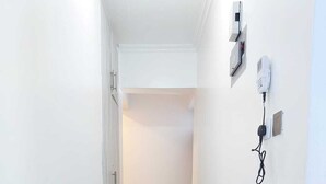 Un couloir étroit au sol carrelé, une porte sur la gauche et un interphone fixé au mur sur la droite.