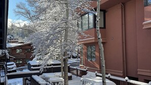 A snow-covered outdoor area with a wooden fence, a small bridge, and a building with large windows. - Ski In, Modern, Cozy Condo on Aspen Mountain - 1 Block from Gondola & Town (Aspen)