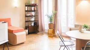 Una habitación con suelo de espiga, un sofá, una mesa de comedor con sillas, una estantería y una ventana con cortinas.