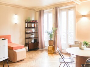 A room with a herringbone floor, a sofa, a dining table with chairs, a bookshelf, and a window with curtains. - Charming apartment with balcony in Paris 11 (Paris)