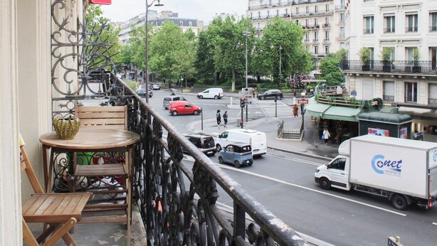 Un balcón con mesa y sillas de madera, con vista a una calle concurrida con coches y edificios.