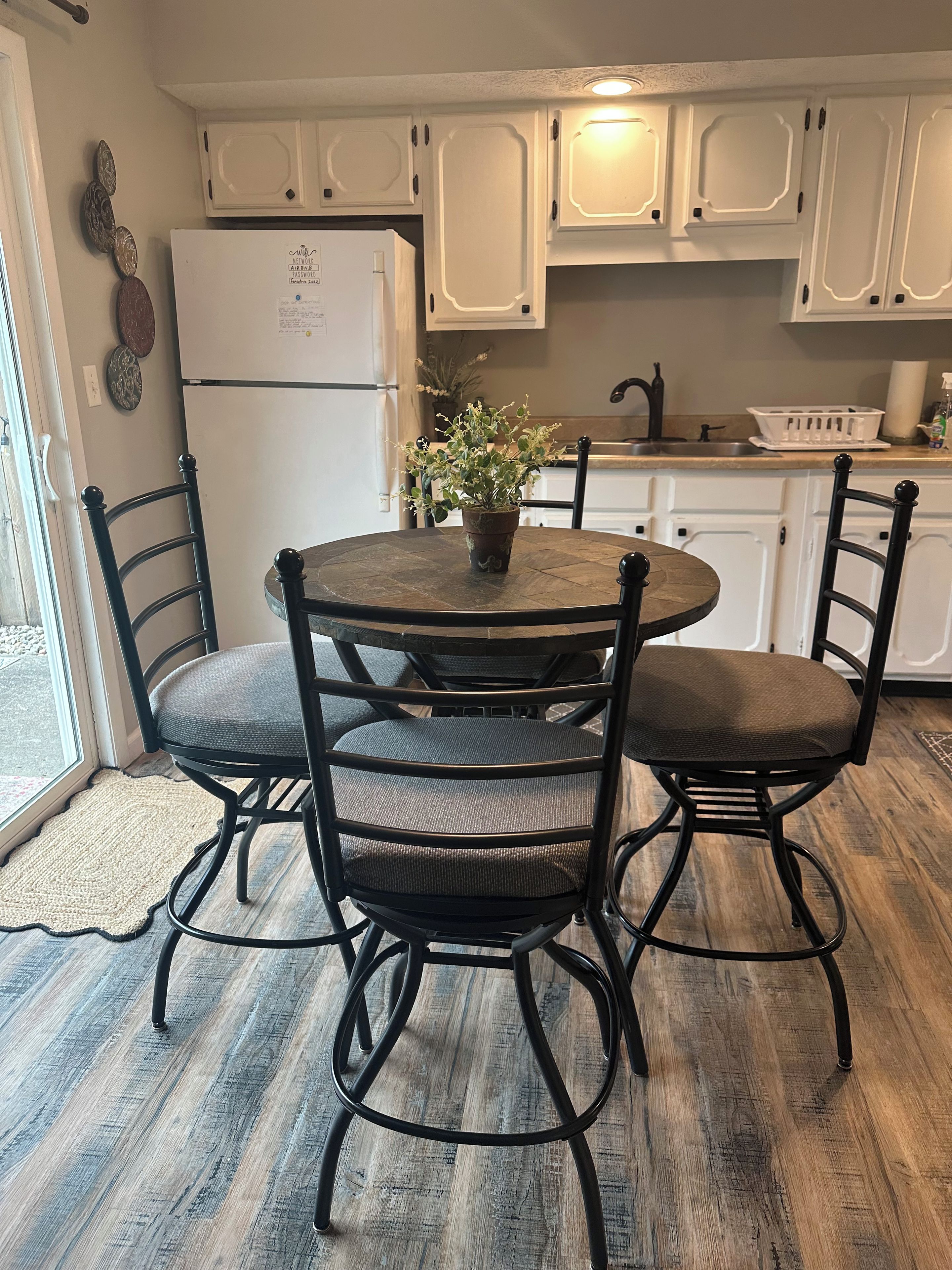 A dining area with a round table and four chairs, a potted plant, and a kitchen in the background.