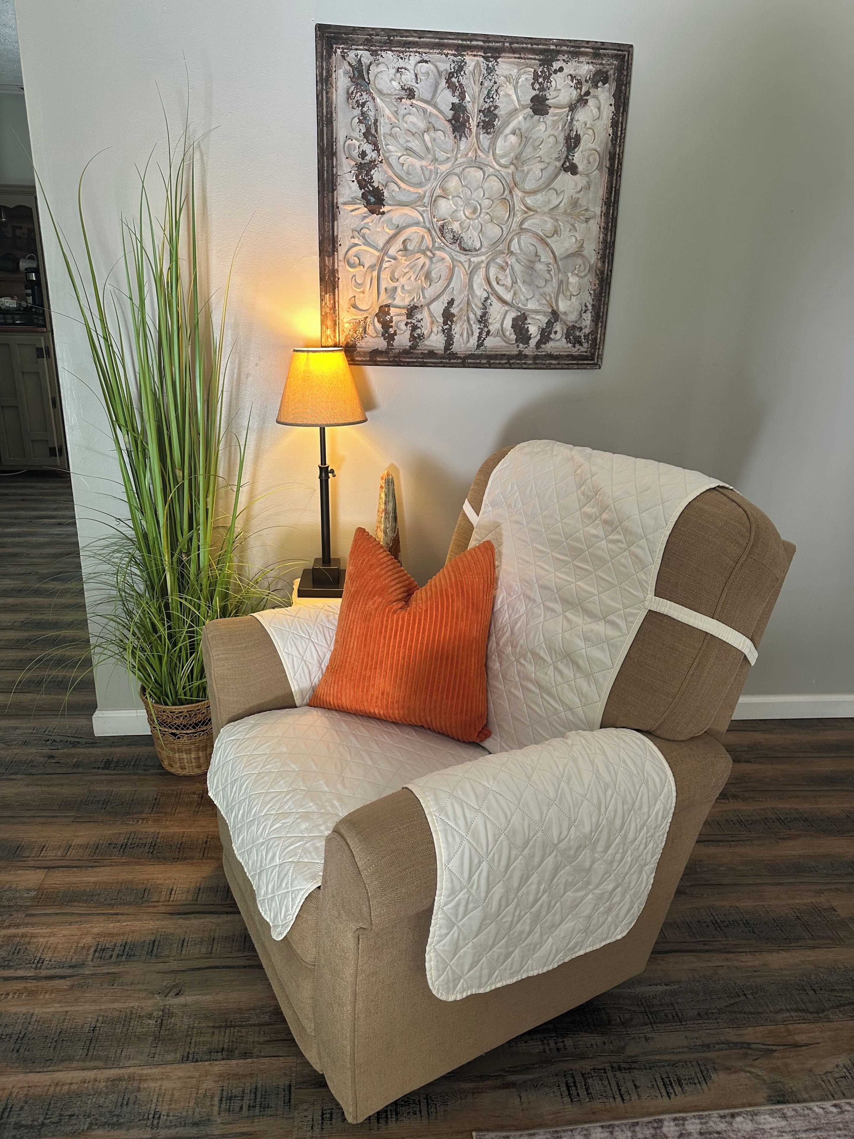 A room with a brown sofa, a white quilted throw, an orange pillow, a potted plant, and a wall-mounted lamp.
