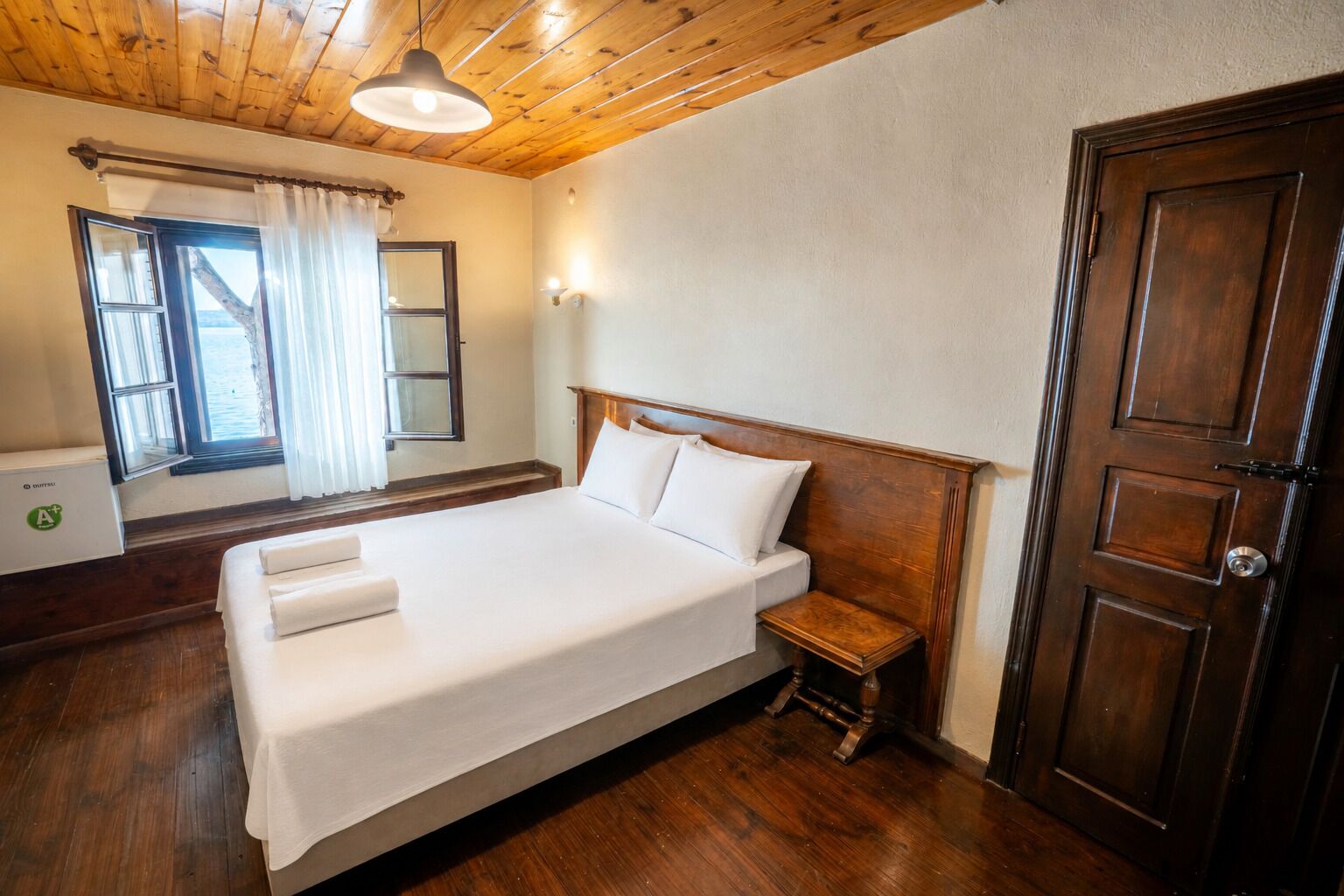 Habitación de hotel con techo de madera, una cama grande con ropa blanca, una mesita de noche, una puerta de madera y una ventana con cortinas.