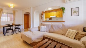 A living room with a sofa, a wooden coffee table, a dining table with chairs, and a kitchen area with wooden cabinets. - 'Nomad House' Apartment with Shared Pool, Wi-Fi, and Air Conditioning (Gran Alacant)