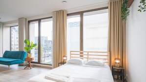 Een hotelkamer met een groot bed, een blauwe stoel, een potplant en uitzicht over de stad.