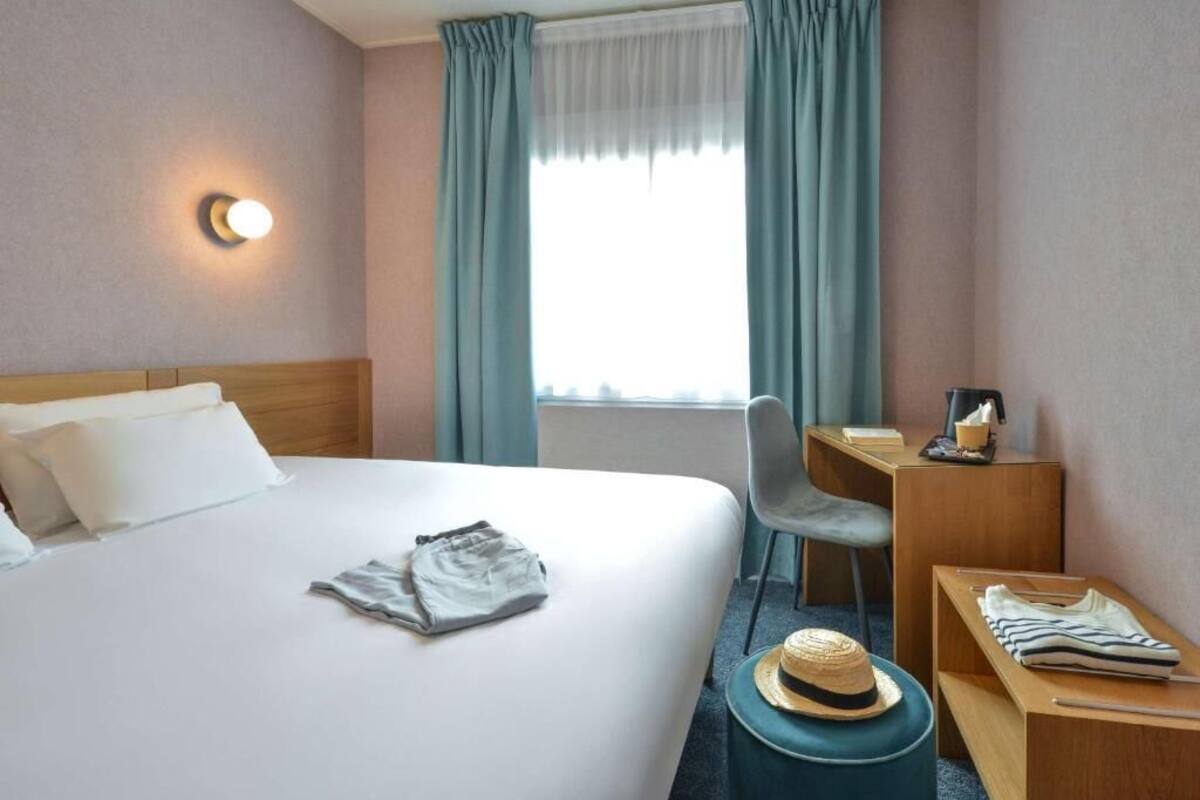Una habitación de hotel con cama, escritorio, silla, mesita y ventana con cortinas.