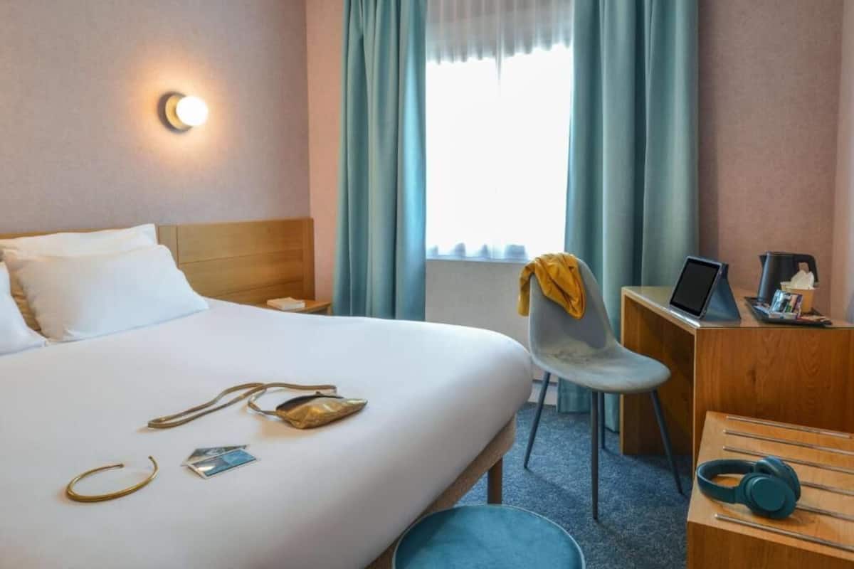 Habitación de hotel con cama, escritorio con computadora portátil, silla, mesita de noche con lámpara y ventana con cortinas.