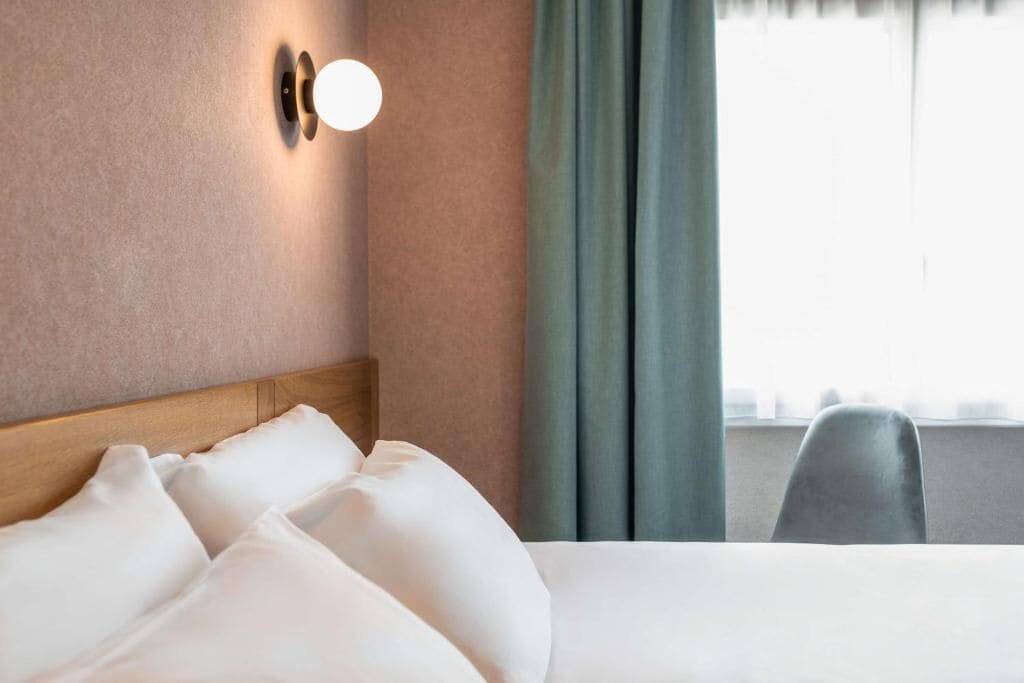 A hotel room with a bed, a headboard, a wall-mounted light, and a window with curtains.