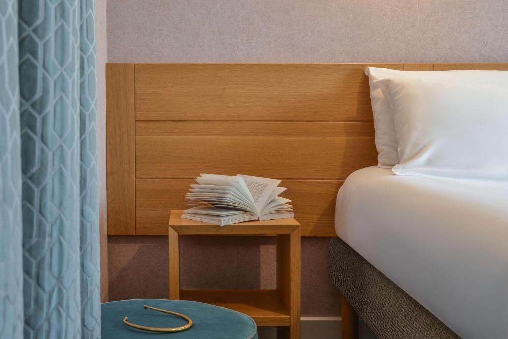 A hotel room with a wooden headboard, a neatly made bed, a small wooden nightstand with an open book, and a blue ottoman.