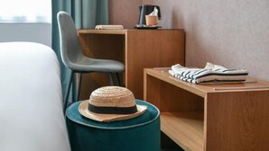 Ein Hotelzimmer mit einem Bett, einem Sessel, einem kleinen Tisch mit einem Hut, einem Regal mit gefalteten Handtüchern und einer Kaffeemaschine.