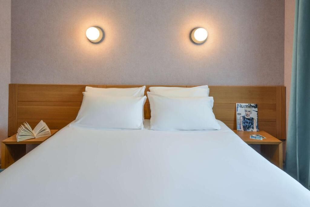 Une chambre d’hôtel avec une tête de lit en bois, deux tables de chevet, un lit avec une literie blanche et deux lampes fixées au mur.
