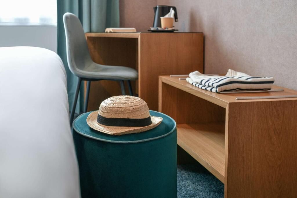 A hotel room with a bed, a chair, a wooden desk, a striped towel, and a straw hat on a round stool.