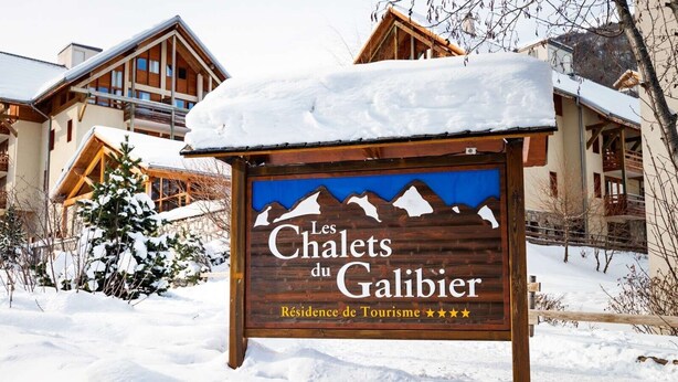 Une enseigne en bois portant les inscriptions « Les Chalets du Galibier » et « Résidence de Tourisme », entourée de neige et de chalets en bois.