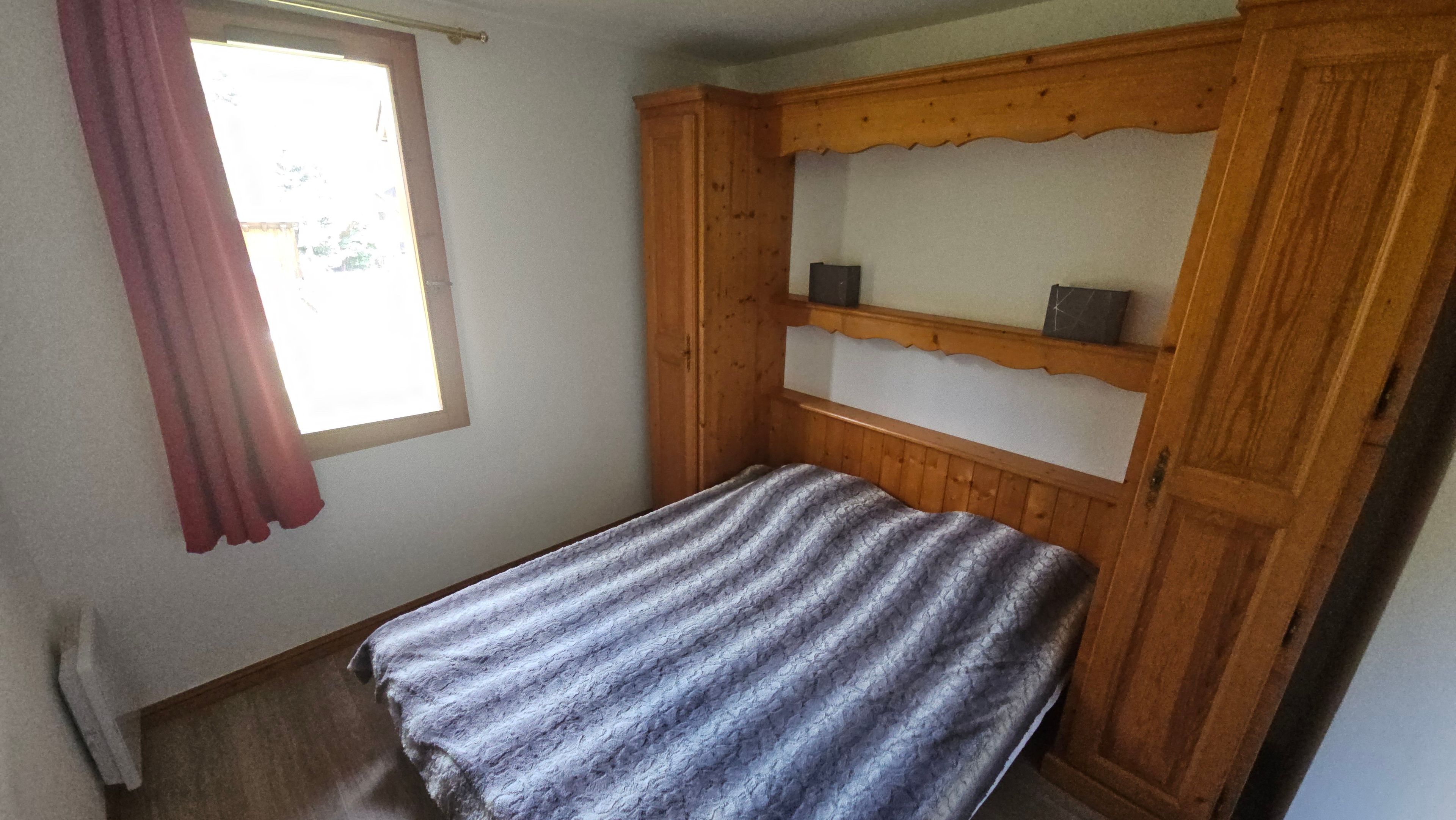Een kleine kamer met een enkel bed, een houten hoofdbord, een raam met gordijn en een vloerbedekking.