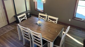 A dining area with a wooden table, white chairs, pendant lights, a bench, and a painting on the wall.