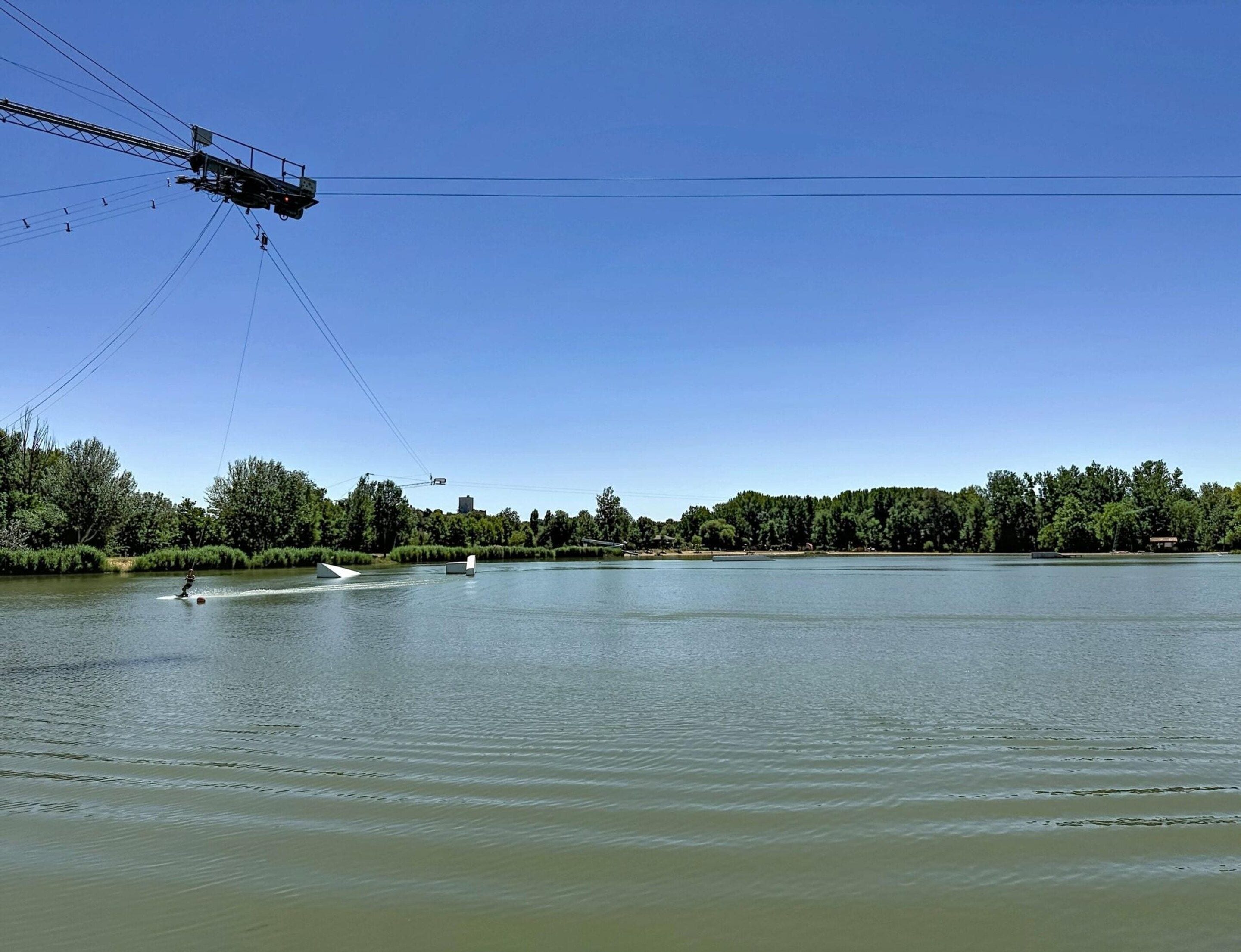 Un lac paisible où une personne fait du wakeboard, entouré d’arbres et sous un ciel dégagé.